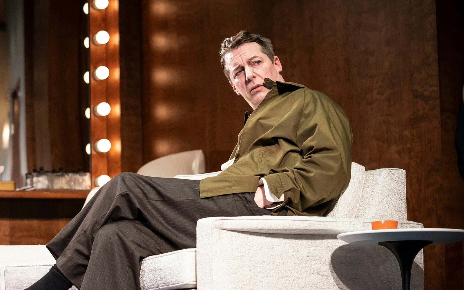 Actor seated on stage set of "Good Night, Oscar" with illuminated mirror in background.