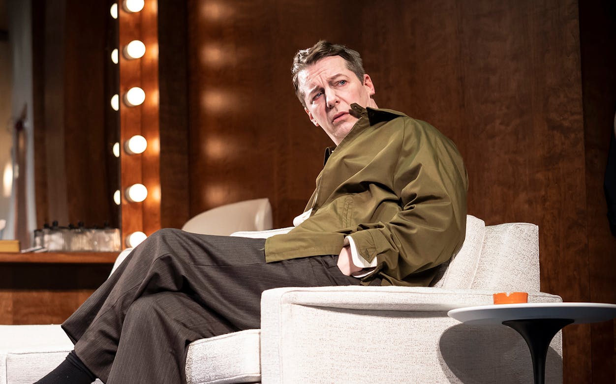 Actor seated on stage set of "Good Night, Oscar" with illuminated mirror in background.