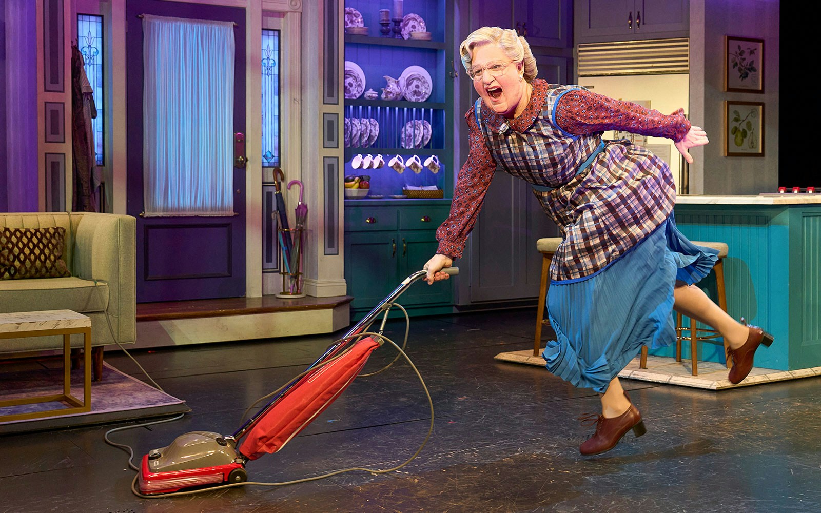 Stage scene of a character dressed as Mrs. Doubtfire vacuuming energetically in a colorful kitchen.