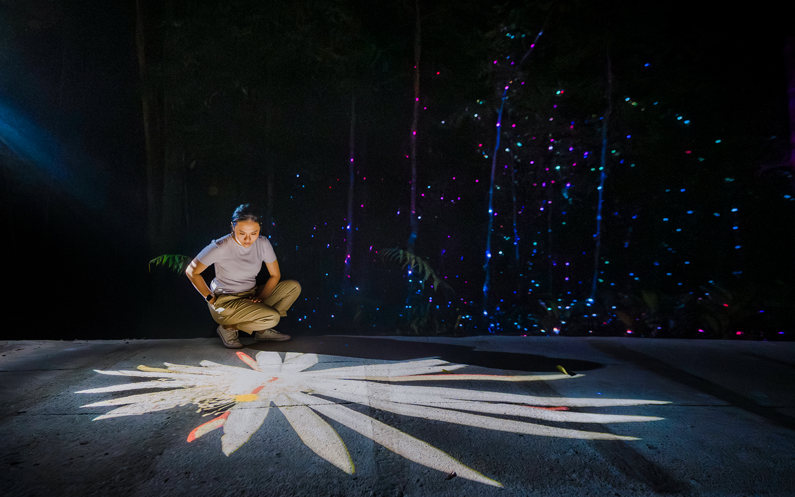 Person interacting with digital projection of a flower, surrounded by colorful lights in a dark setting.