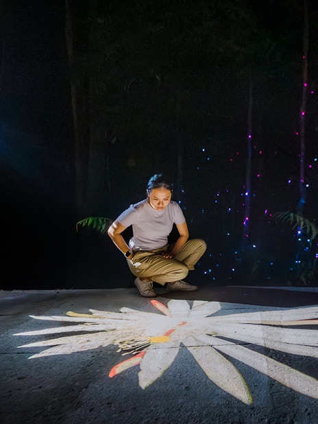 Person interacting with digital projection of a flower, surrounded by colorful lights in a dark setting.