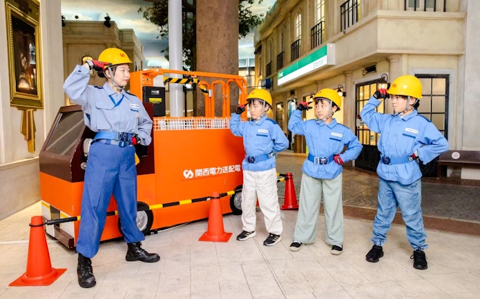 Municipal workers at KidZania Osaka Koshien in uniforms and helmets near an orange vehicle.