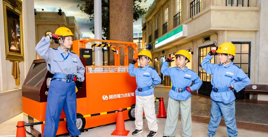 Municipal workers at KidZania Osaka Koshien in uniforms and helmets near an orange vehicle.