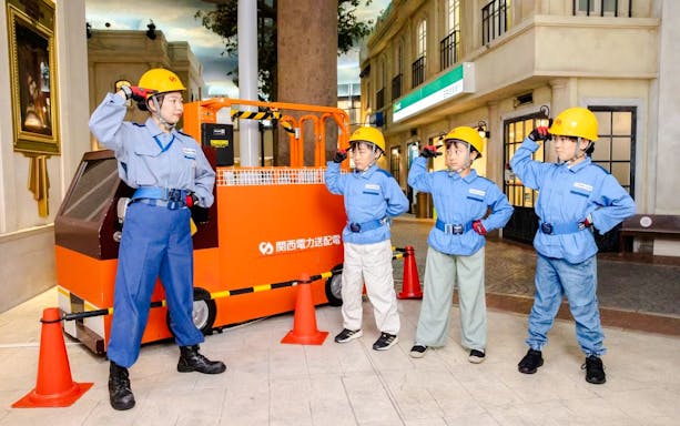 Municipal workers at KidZania Osaka Koshien in uniforms and helmets near an orange vehicle.