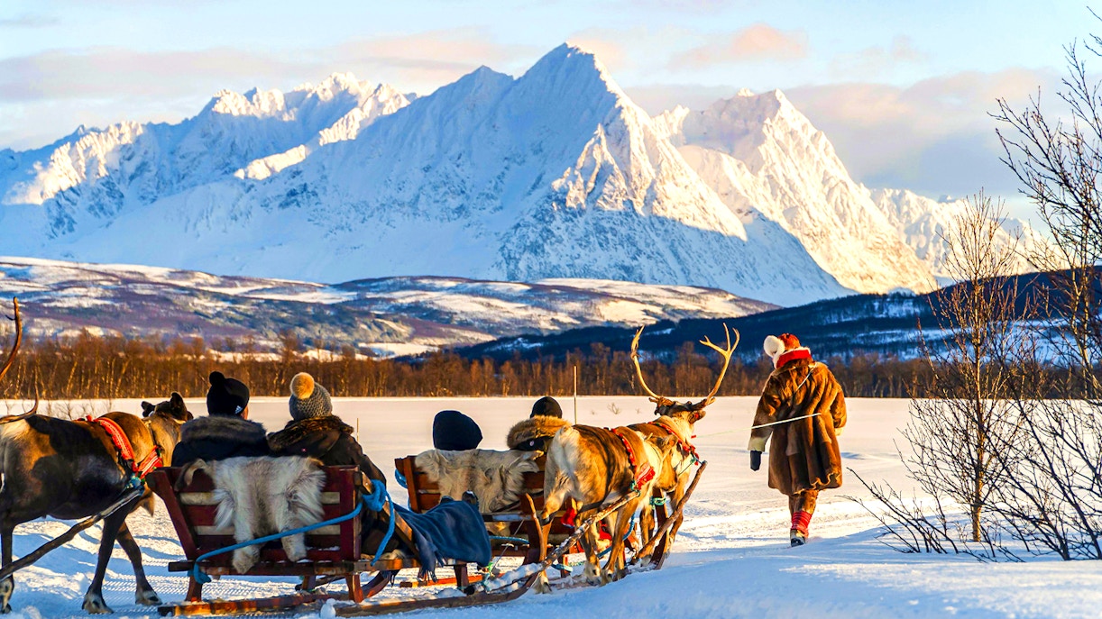 Tourists on a reindeer sled in Tromso, guided by a Sami person, with snowy mountains in the background.