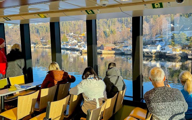 Sightseeing cruise passengers view Oslo fjord landscape through large windows.