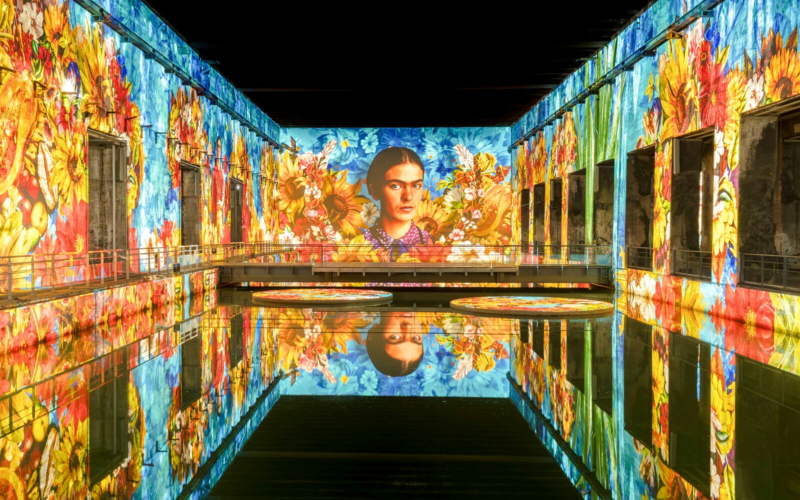 Frida Kahlo artwork projected at Bassins Des Lumieres exhibition, vibrant floral display.
