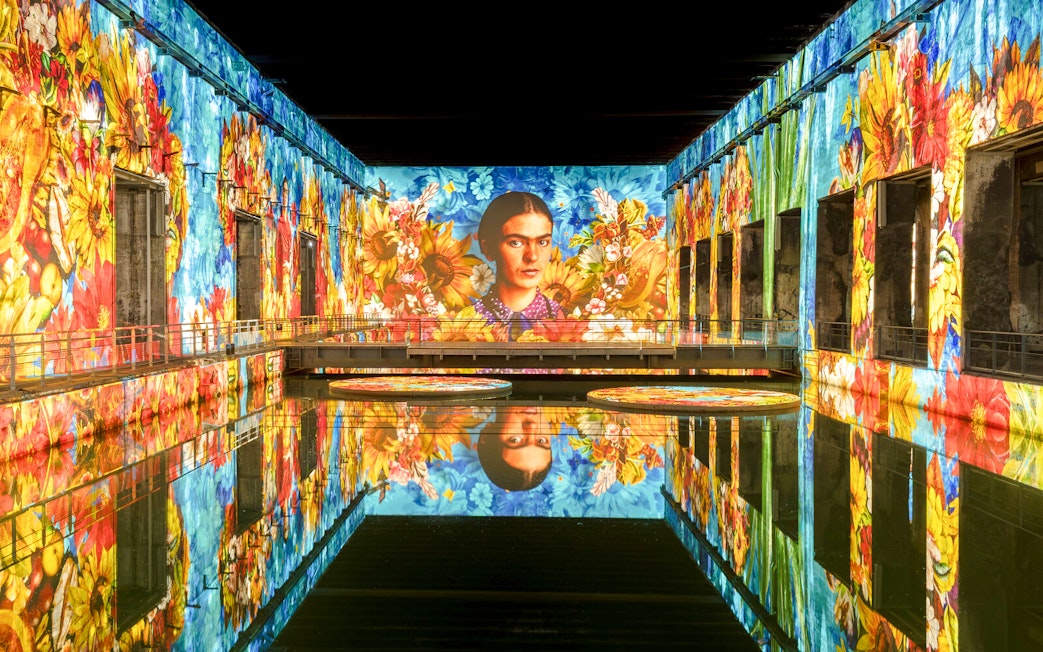 Frida Kahlo artwork projected at Bassins Des Lumieres exhibition, vibrant floral display.