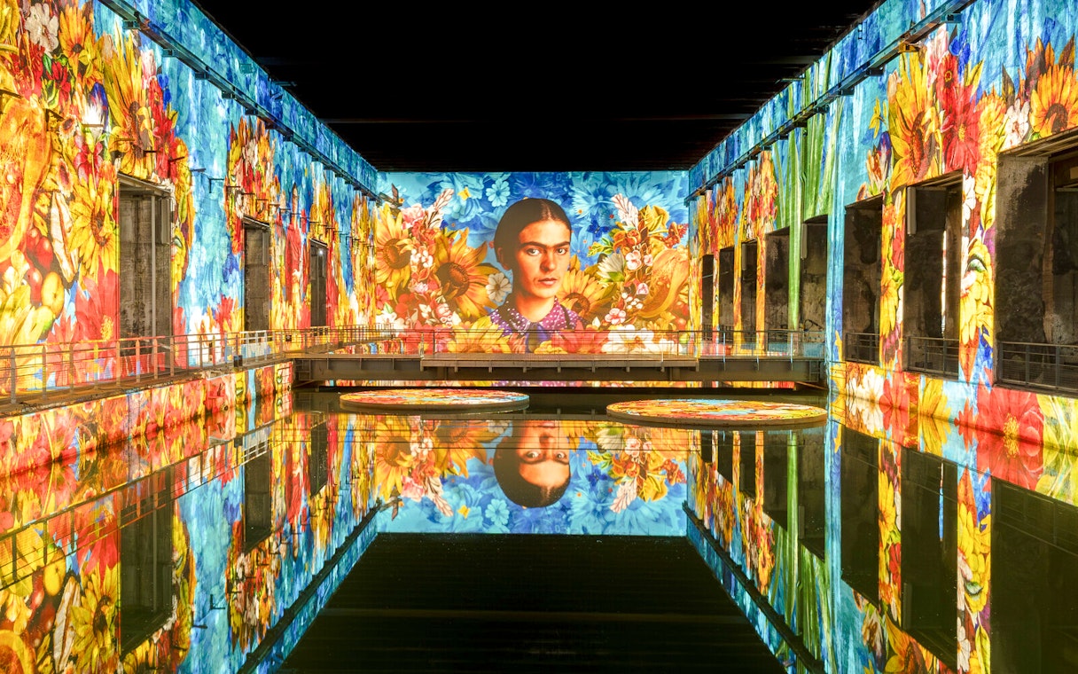 Frida Kahlo artwork projected at Bassins Des Lumieres exhibition, vibrant floral display.