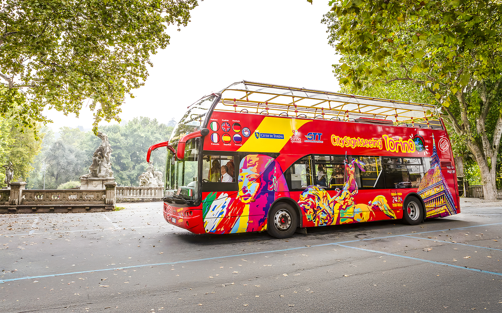 Turin hop-on hop-off bus near a park with statues and trees.
