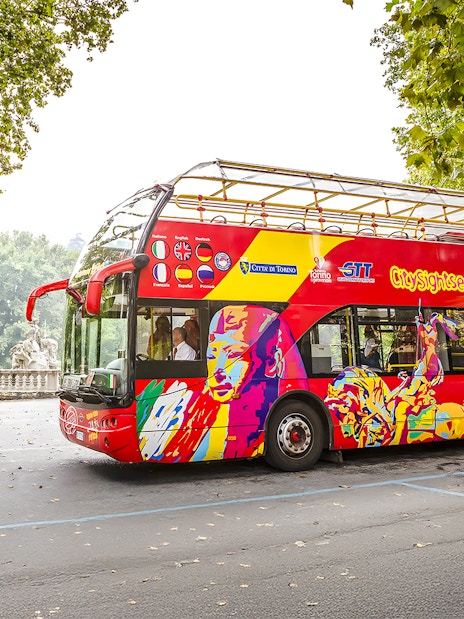 Turin hop-on hop-off bus near a park with statues and trees.