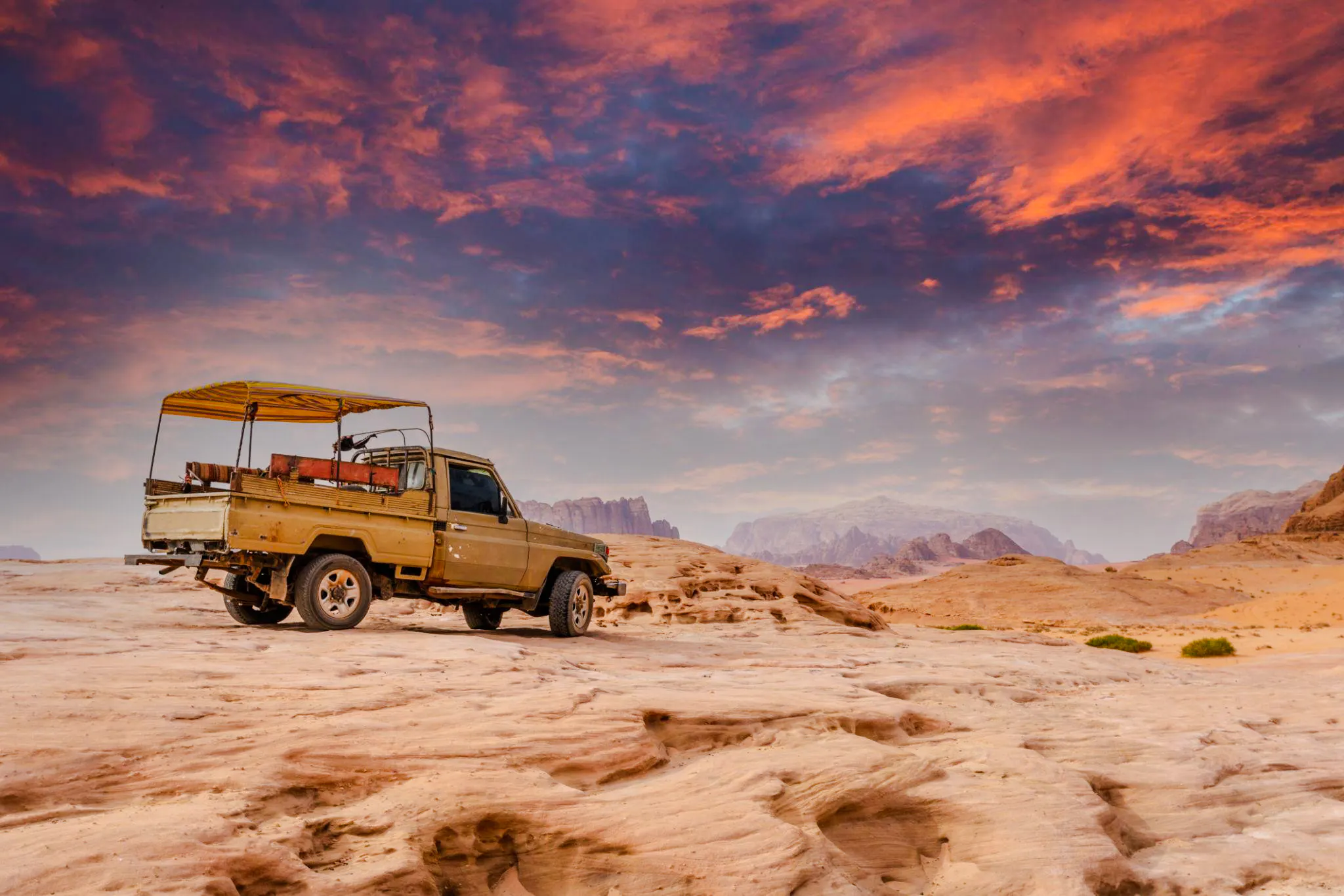 Jeep on rocky terrain at sunset in Wadi Rum, Jordan, with mountains in the background.