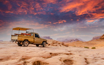 Jeep on rocky terrain at sunset in Wadi Rum, Jordan, with mountains in the background.