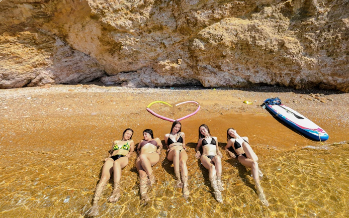 Five women relaxing on a sandy beach, partially submerged in clear water, with a paddleboard nearby.