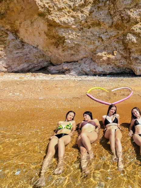 Five women relaxing on a sandy beach, partially submerged in clear water, with a paddleboard nearby.