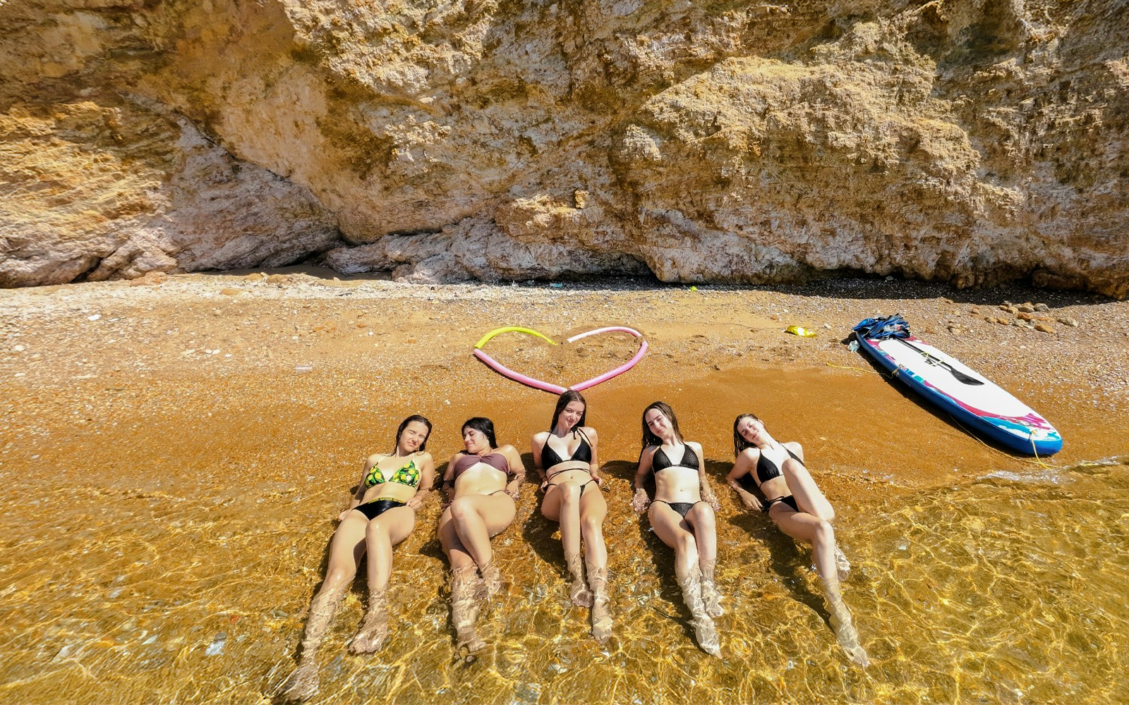 Five women relaxing on a sandy beach, partially submerged in clear water along the Athenian coastline.