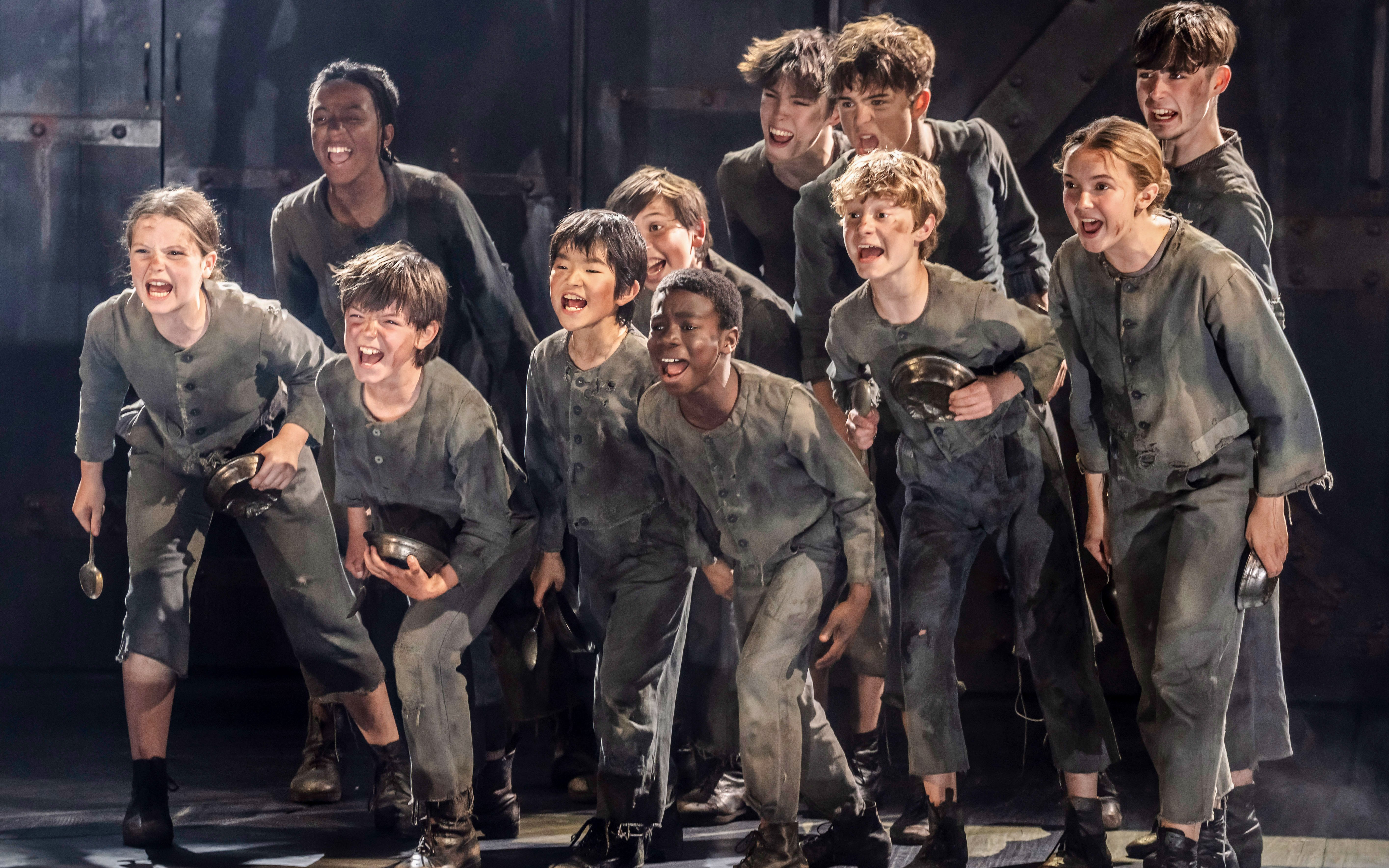 Children in ragged clothing holding bowls, performing a scene from "Oliver" musical production.