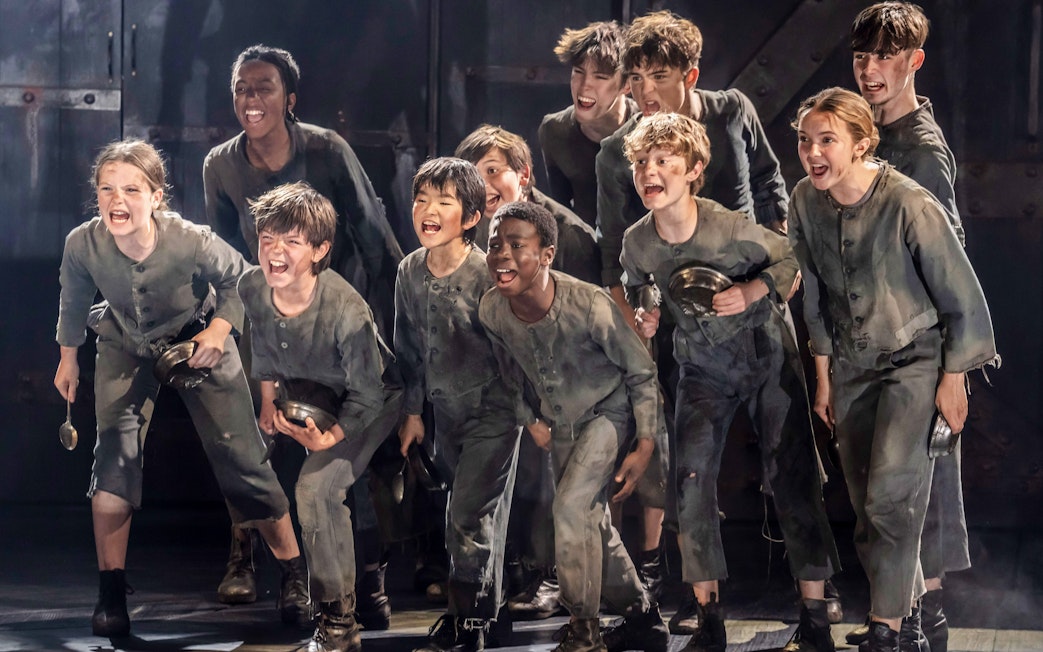 Children in ragged clothing holding bowls, performing a scene from "Oliver" musical production.