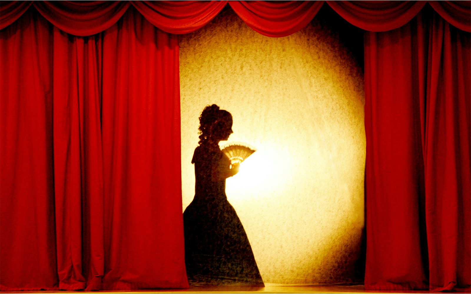 Shadow of a woman holding a fan on stage with red curtains in a theater.