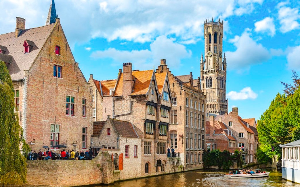 Canal in Bruges, Belgium with Belfry of Bruges in background and boat on water.