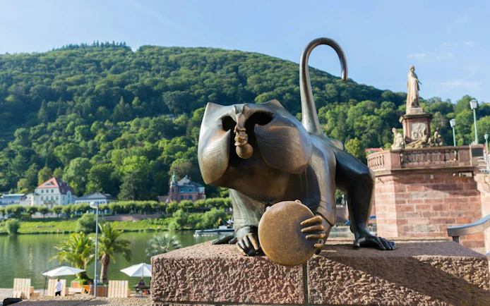 Heidelberg Bridge Monkey statue with scenic hillside backdrop.