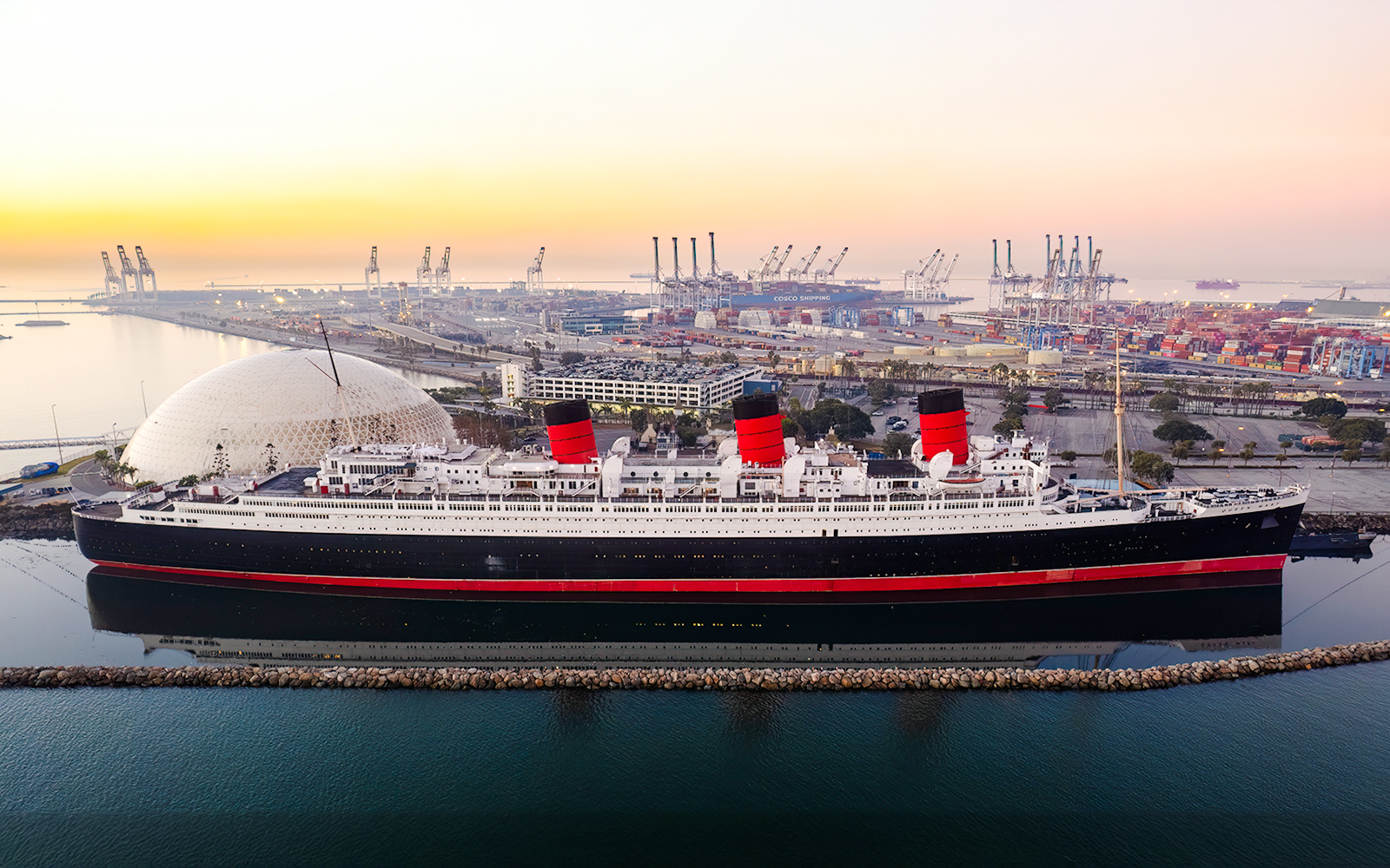 Queen Mary ship docked at sunrise with port cranes in the background.