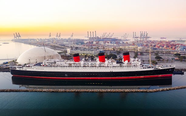 Queen Mary ship docked at sunrise with port cranes in the background.