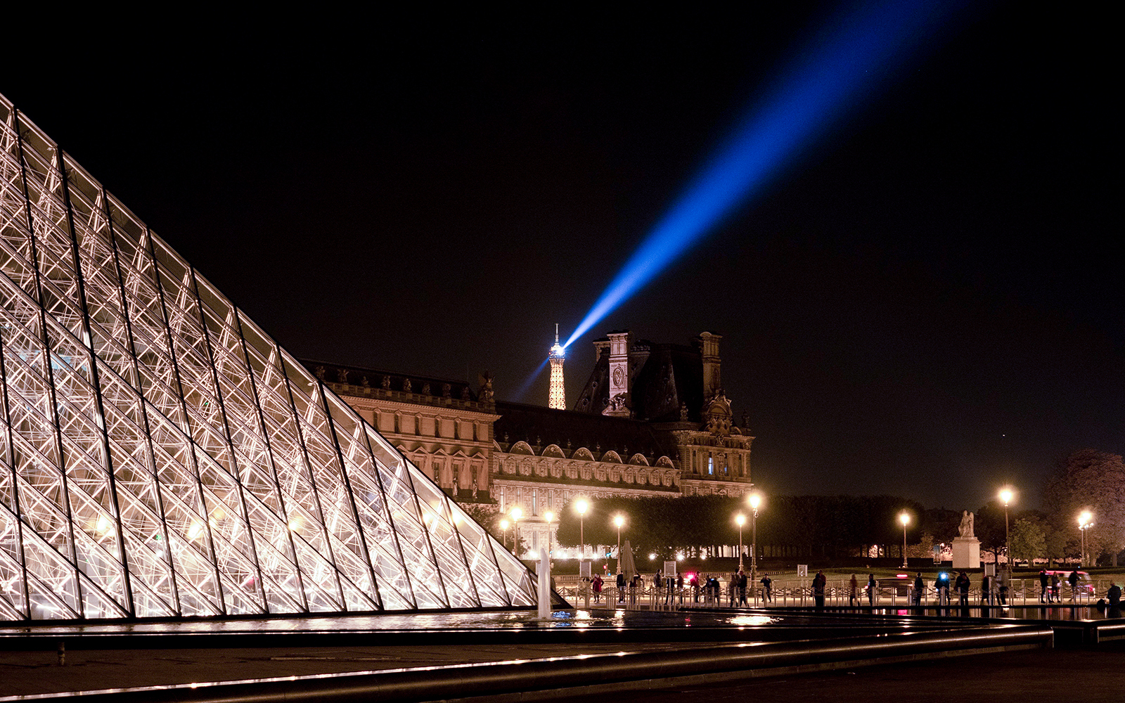 Louvre Pyramid illuminated at night with Eiffel Tower spotlight in Paris.