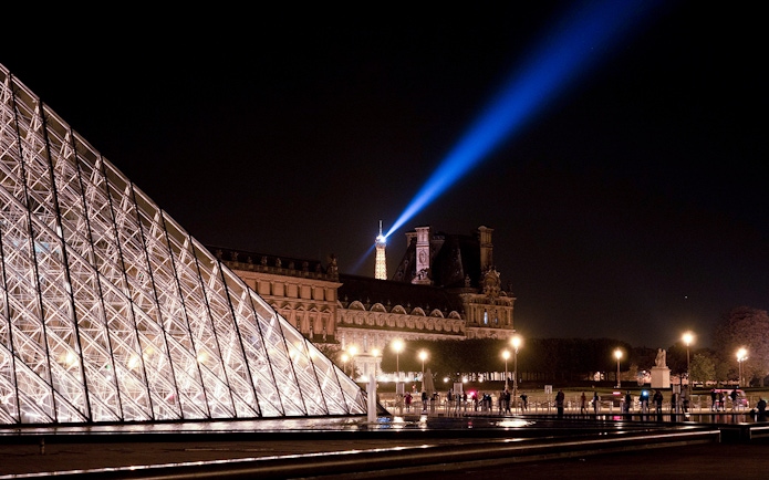 Louvre Pyramid illuminated at night with Eiffel Tower spotlight in Paris.