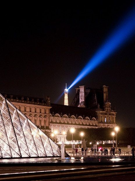 Louvre Pyramid illuminated at night with Eiffel Tower spotlight in Paris.
