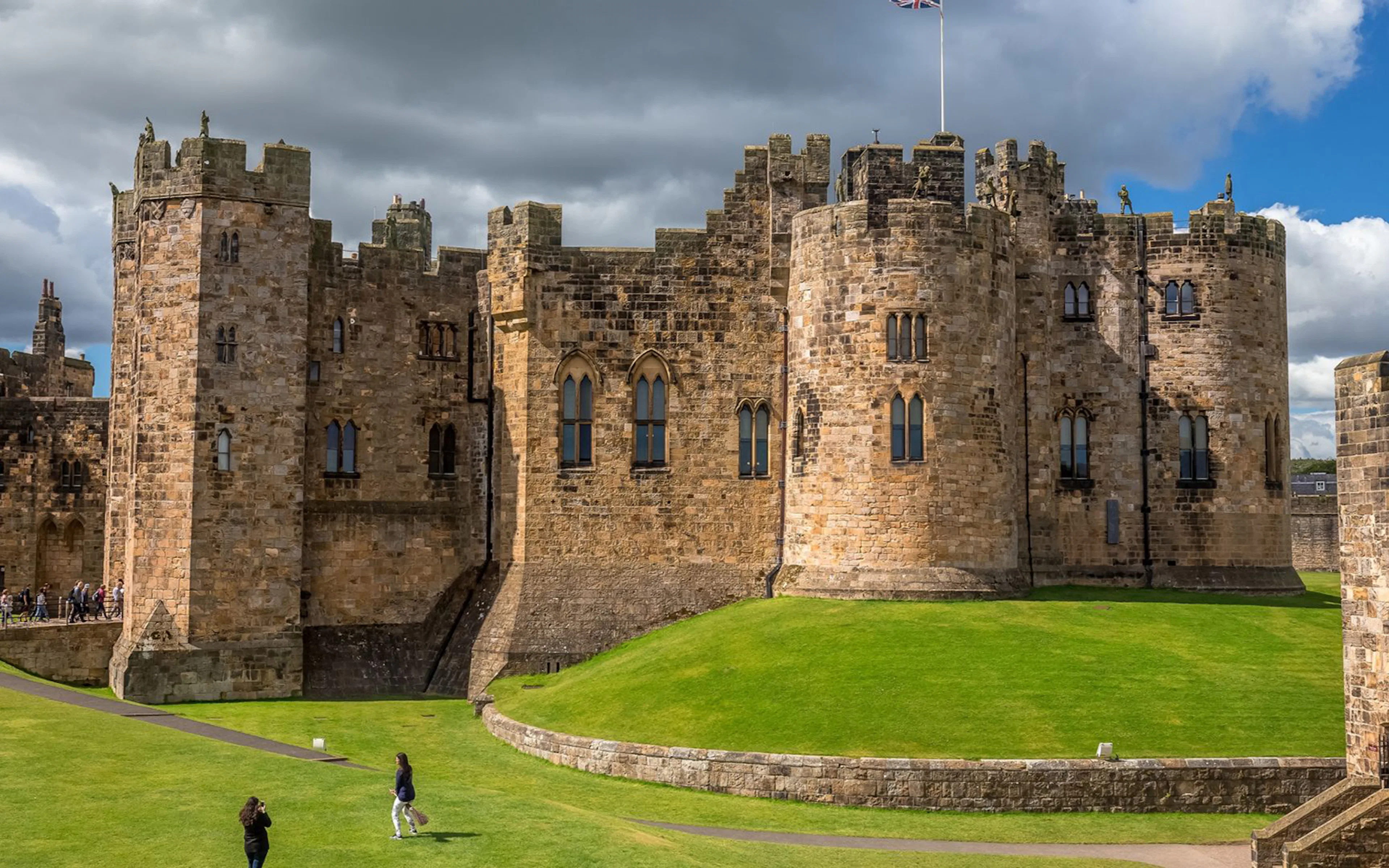 Alnwick Castle exterior with stone towers and green lawn under cloudy sky.