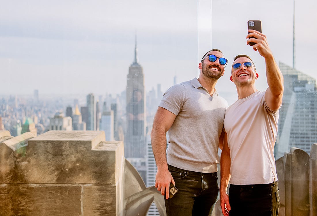 Men taking selfie at Top of the Rock, Rockefeller Center, New York City skyline in background.
