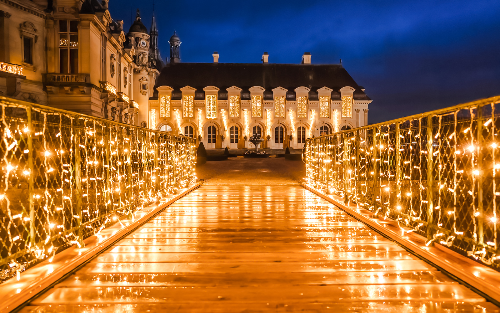 Chateau of Chantilly illuminated at night with festive lights on a bridge.