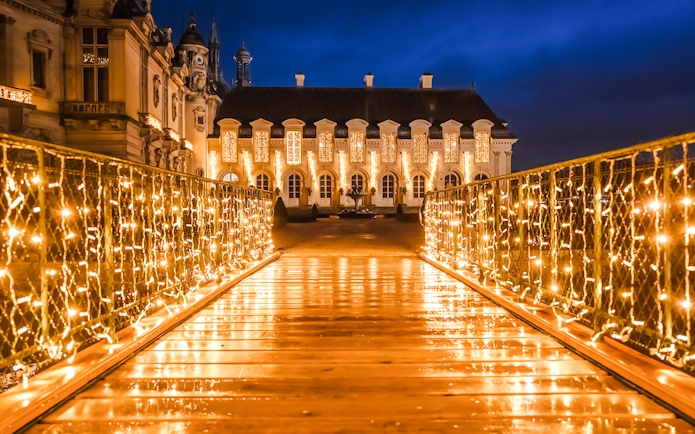 Chateau of Chantilly illuminated at night with festive lights on a bridge.