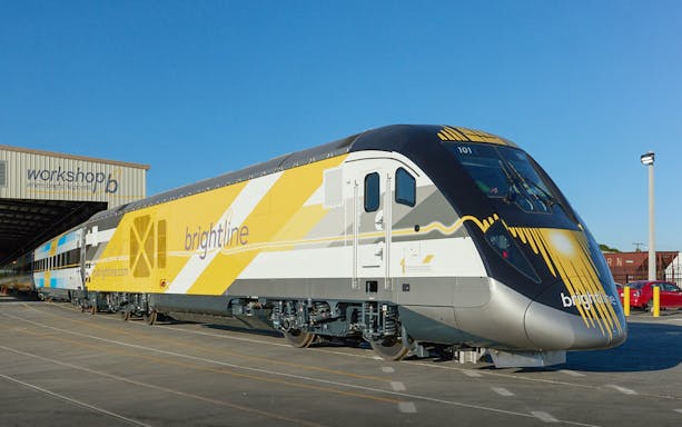 Brightline train parked outside a workshop under clear blue sky.