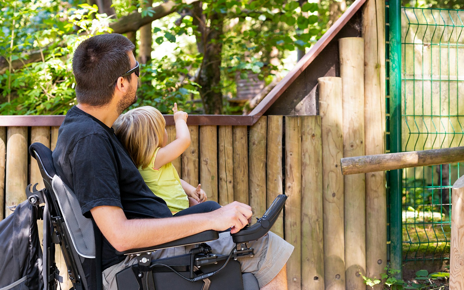 Man in wheelchair with child pointing at animals in a zoo enclosure.