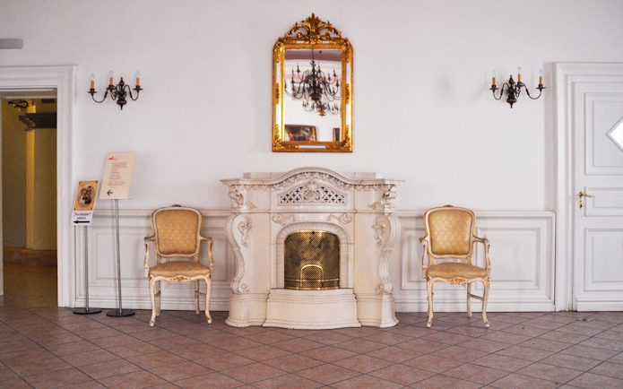 Gödöllő Royal Palace interior with ornate fireplace and chairs.