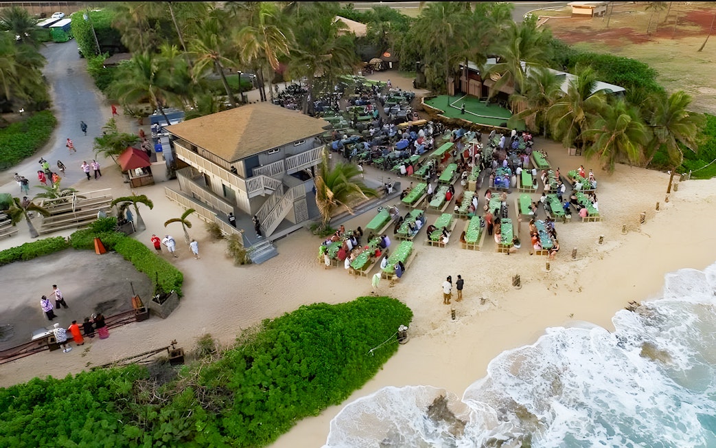 Drone view of Germaine's Luau with guests seated near the shore in Oahu, Hawaii.