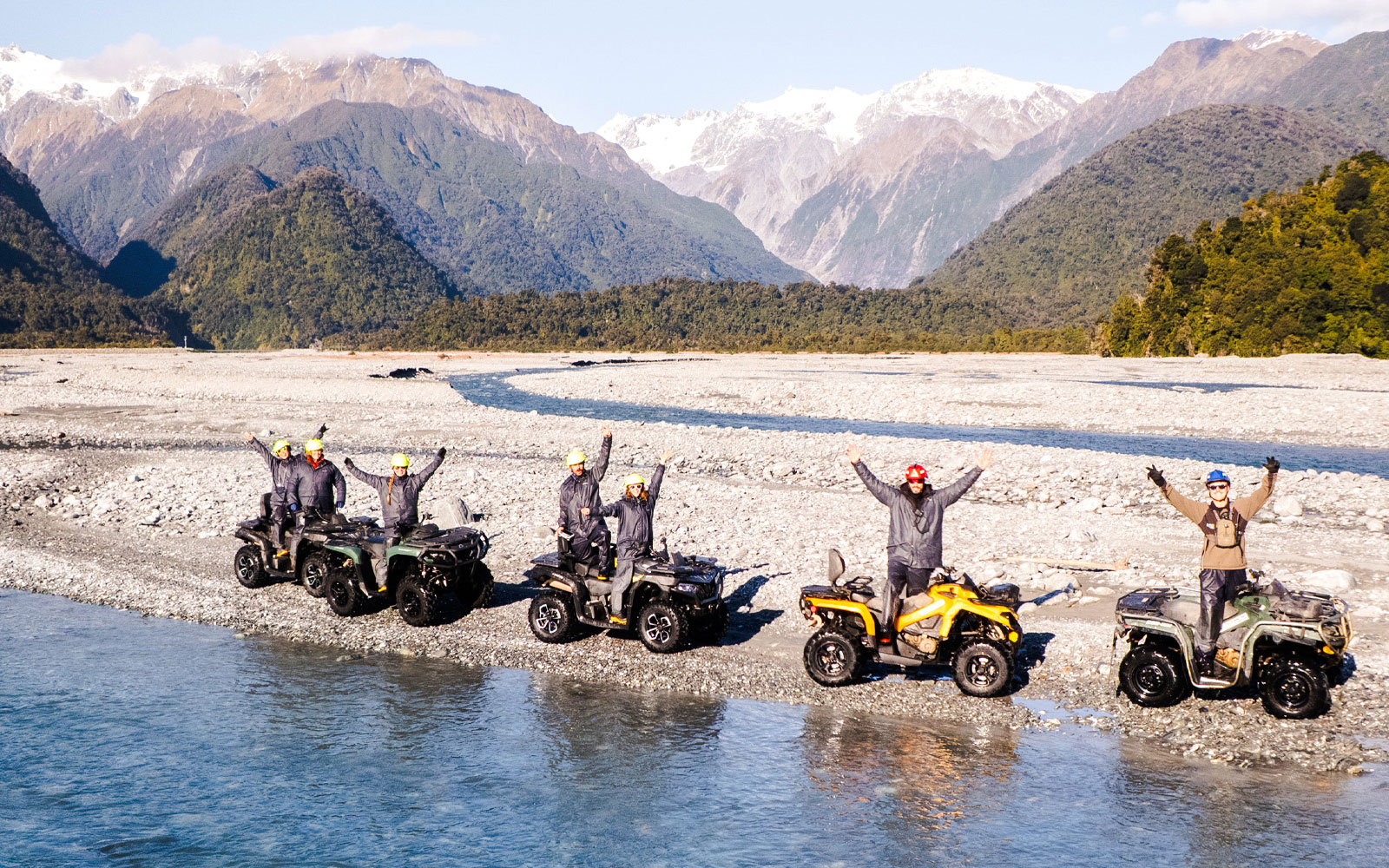Group quad biking by river at Franz Josef, New Zealand with mountain backdrop.