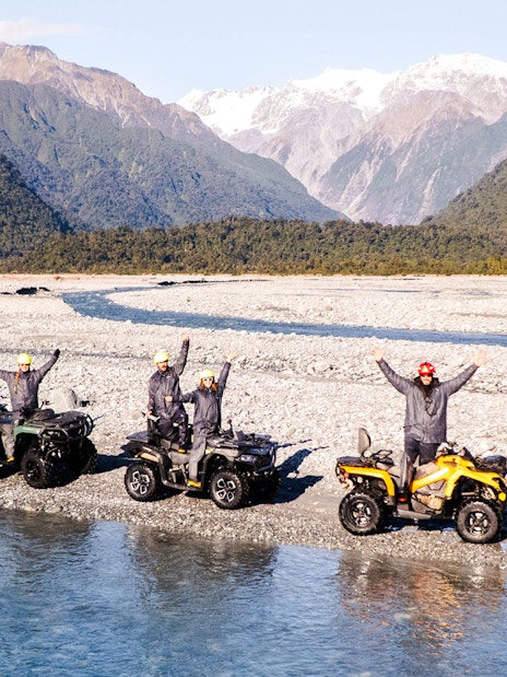 Group quad biking by river at Franz Josef, New Zealand with mountain backdrop.