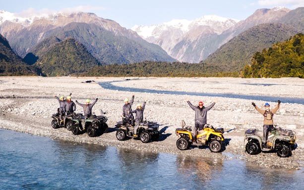 Group quad biking by river at Franz Josef, New Zealand with mountain backdrop.