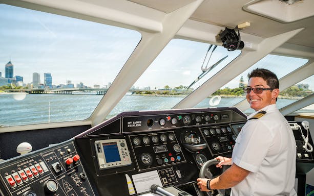 Pilot steering Fremantle Swan River Scenic Cruise with Perth skyline in view.