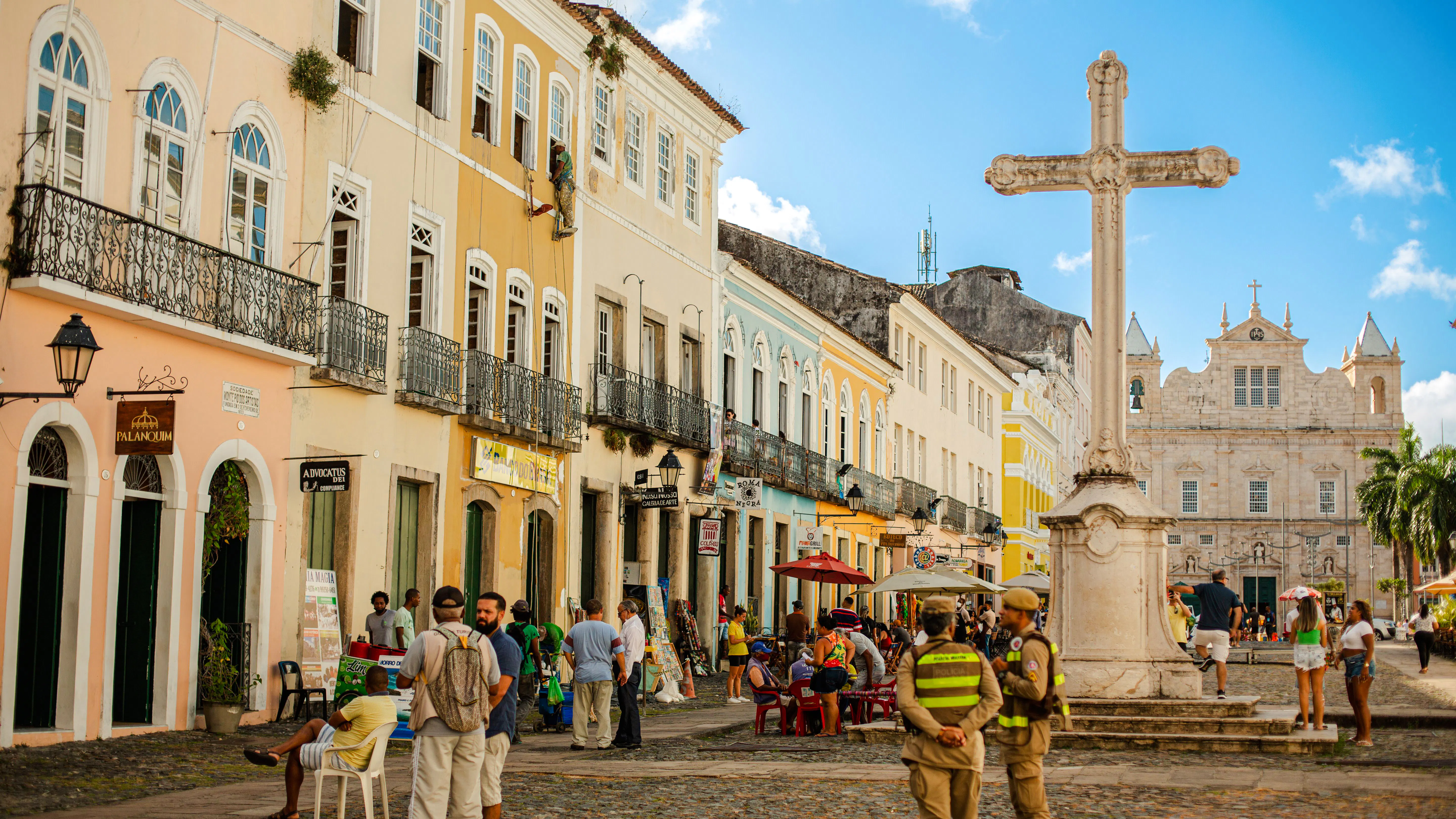 Colorful colonial buildings and a stone cross in Pelourinho, Salvador, with people walking and socializing.