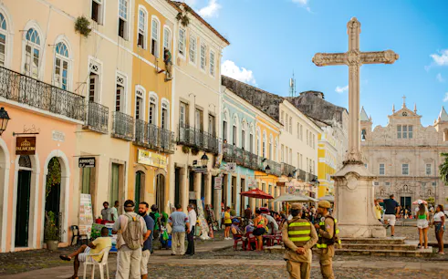Colorful colonial buildings and a stone cross in Pelourinho, Salvador, with people walking and socializing.