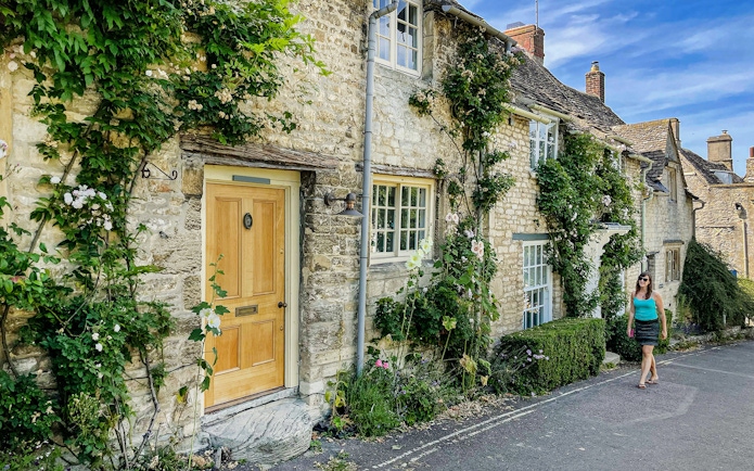 Cotswolds stone cottage with climbing plants and a person walking nearby.