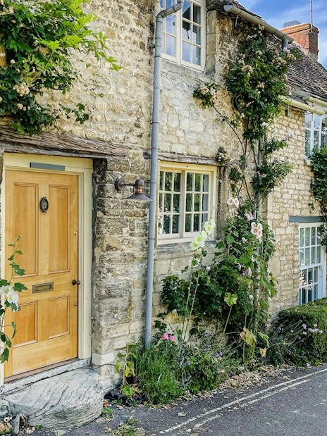 Cotswolds stone cottage with climbing plants and a person walking nearby.