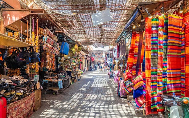 Marrakech market with colorful textiles and sandals displayed, part of Agadir full-day city tour.