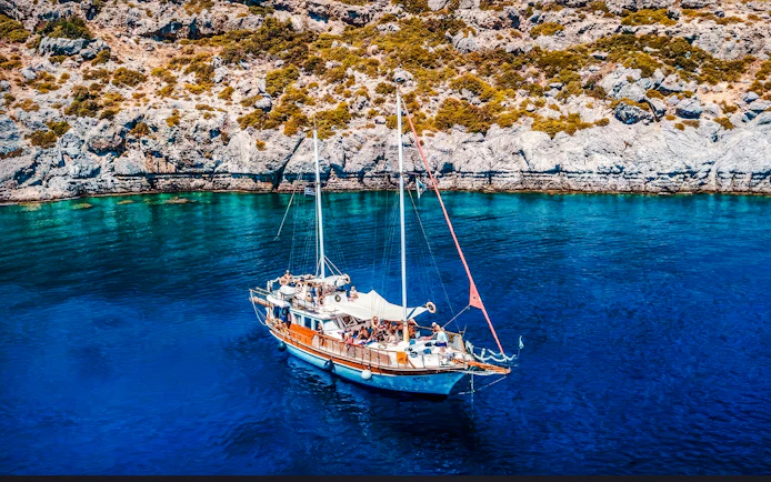 Sailing boat on clear blue waters near rocky coast, Rhodes All-Inclusive Cruise.