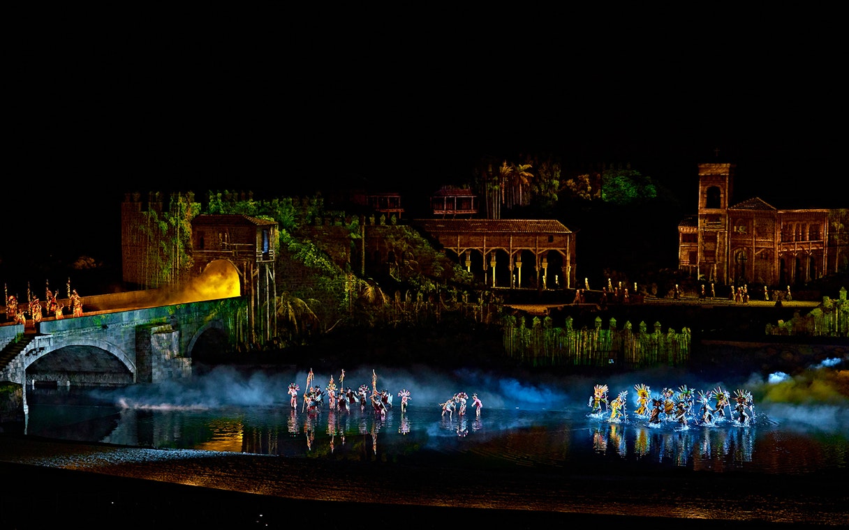 Nighttime performance at Puy du Fou España with dancers in colorful costumes on water stage.