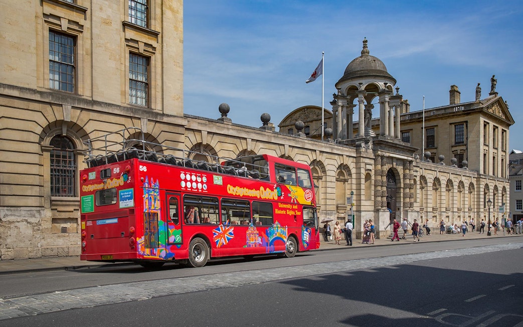 Red double-decker bus on Oxford street near historic building, part of Oxford by Rail day tour from London.